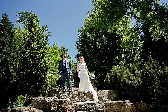 Couple portrait of bride and groom standing on stone steps, bride holding white bouquet in sleeveless gown with long train, blue sky behind