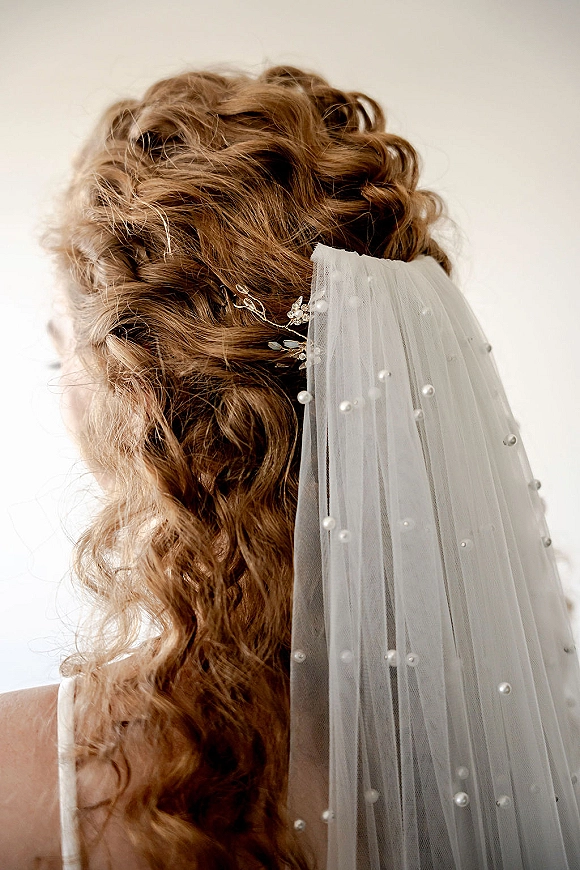 Bridal hairstyle in a wedding updo with soft curls under a pearl veil, shown from the back against a clean white wall backdrop