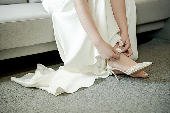 Bridal shoes lace wedding heels with ankle strap buckle beside the bridal gown hem, resting on a sofa over a carpeted floor