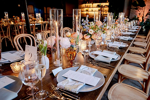 Reception tablescape with long banquet table setting, taper candles in glass hurricanes, floral centerpieces and amber goblets in a dim indoor space