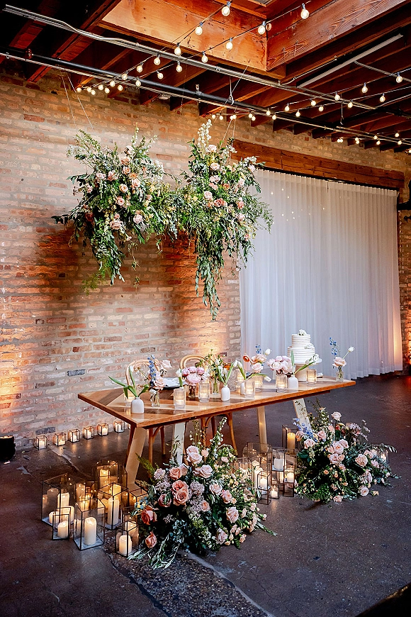 Reception sweetheart table with hanging floral installation, blush roses, candlelit lanterns, and white cake against an exposed brick wall backdrop