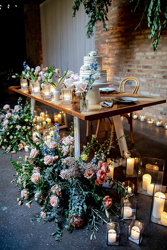 Wedding cake table with a tiered buttercream wedding cake on a stand, roses and blue blooms, candles and black lanterns against a brick wall backdrop