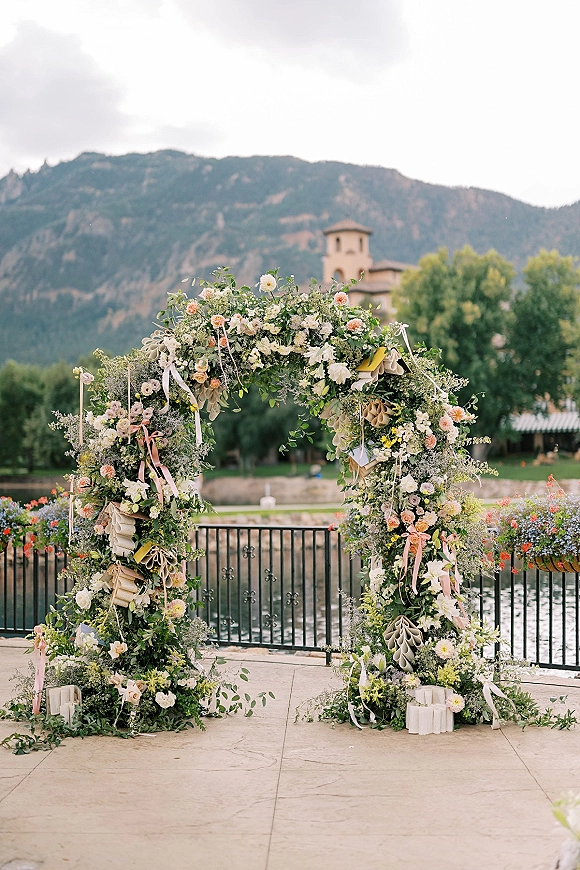 Wedding ceremony arch with pastel florals and greenery garland on a stone patio, with pillar candles and mountain river backdrop