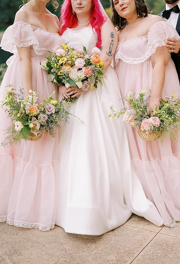 Bridesmaid portrait with bride and blush bridesmaid dresses, holding colorful wildflower bouquets on stone pavement amid outdoor greenery