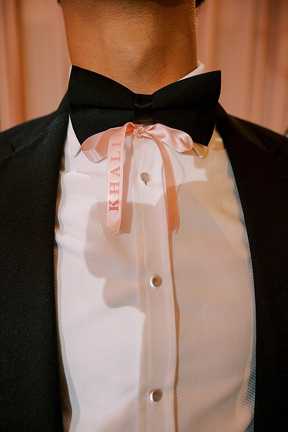 Groom boutonniere alternative with personalized ribbon boutonniere, pink satin bow on black tuxedo lapel and bow tie against pink curtain backdrop