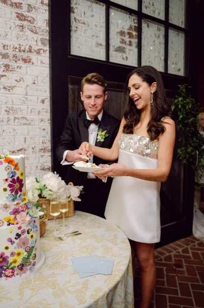 Wedding cake cutting as bride in a strapless mini dress and groom in tux slice a floral three-tier cake by a black door and brick wall