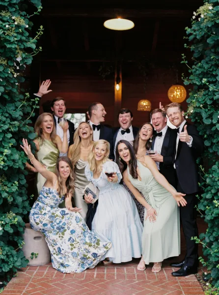 Wedding group photo of bridesmaids and groomsmen cheering with wine glasses under a greenery arch by an ivy-covered wooden doorway on a brick patio