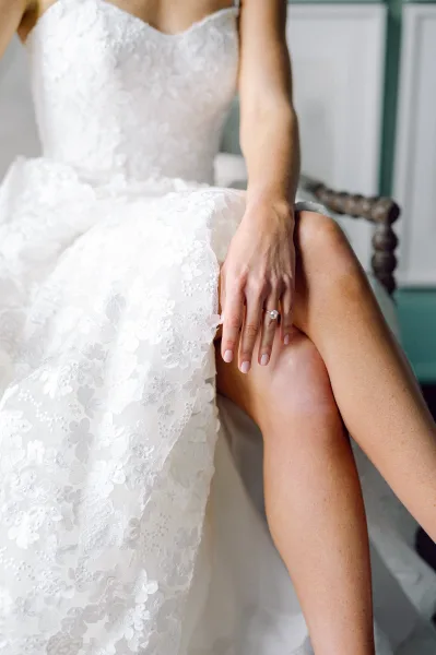 Engagement ring diamond solitaire ring on bride’s pale pink manicured hand, lace wedding dress detail visible as she sits indoors