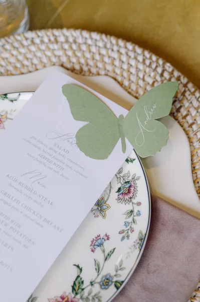 Wedding place card and butterfly place card with calligraphy on a floral china plate, paired with menu card, napkin, and woven charger