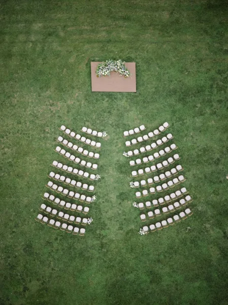 Ceremony setup with outdoor wedding ceremony chairs in a curved split-aisle layout, floral arch on a wooden platform on grass lawn