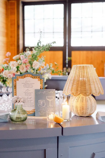 Wedding bar decor with signature cocktail sign on the counter, lit by candles beside a wicker table lamp and floral buds by big windows
