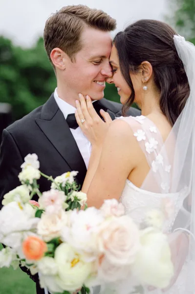 Wedding couple portrait with bride and groom close up, touching foreheads as she holds a soft pastel bouquet on a green lawn with trees
