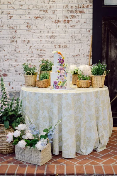 Wedding cake with pressed flower wedding cake design on a round table with patterned cloth, set against a white brick wall and door