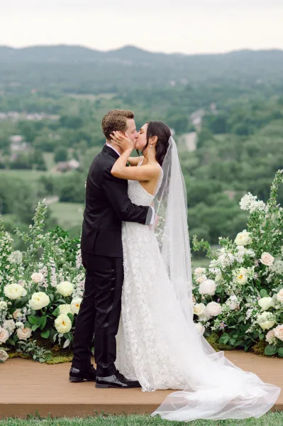 Wedding kiss portrait of bride and groom kissing, long veil and lace dress on a floral-lined terrace with mountain valley view behind