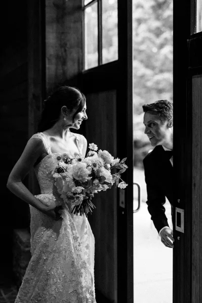 First look moment as bride enters the doorway holding a bouquet, facing groom in suit by a wood wall and bright windows with trees beyond