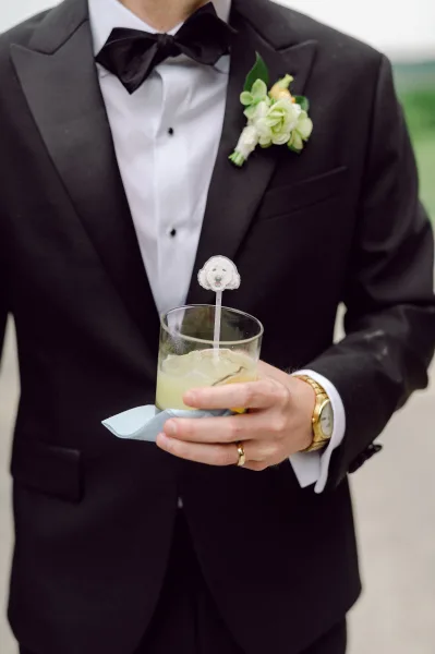Groom portrait in a black tuxedo groom look, holding a cocktail glass with stirrer, boutonniere on lapel against blurred greenery