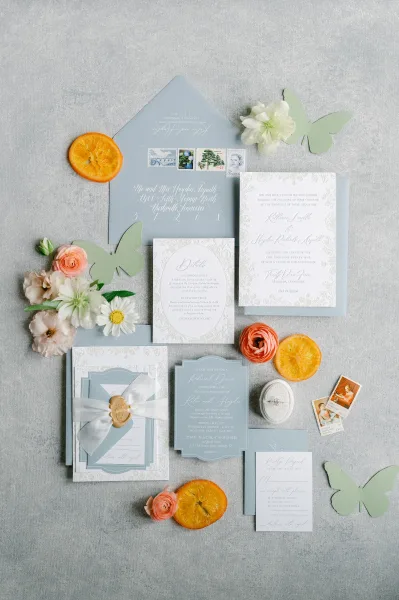 Invitation suite wedding invitation flat lay with dusty blue cards, wax seal and ribbon, ring, flowers, butterflies, and orange slices on gray linen