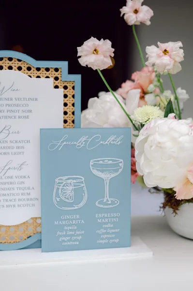 Wedding bar menu on blue cardstock with calligraphy and cocktail icons, propped on rattan backing beside pastel peonies on a white table