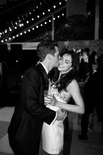 Wedding kiss portrait of bride and groom dancing as he kisses her cheek, string lights glowing over an outdoor night reception dance floor