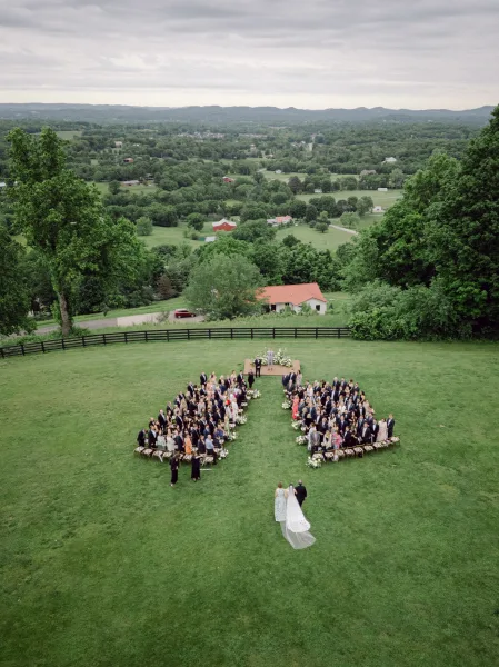 Outdoor wedding ceremony seen in an aerial wedding ceremony view, with circular chairs, aisle flowers, and arch on a grassy lawn under clouds
