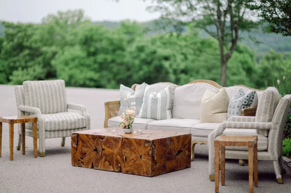 Wedding lounge seating with an outdoor sofa, upholstered chairs and pillows around a carved wood coffee table on a patio with hills beyond
