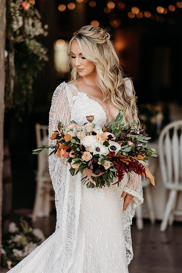 Bridal portrait of a bride holding bouquet in a long-sleeve lace gown with hair pins, looking down beneath barn string lights and chairs