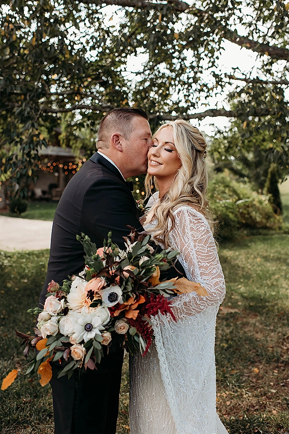 Couple portrait of bride and groom embrace as he kisses her cheek, bouquet and lace sleeves under trees with string lights in a garden