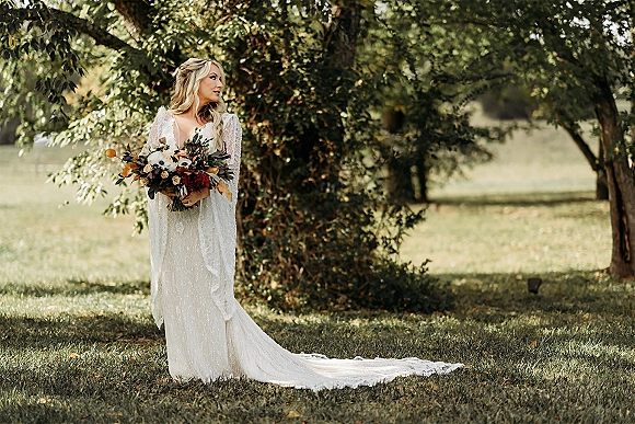 Bridal portrait of a bride holding bouquet of roses and greenery, wearing a long-sleeve lace gown with train in a sunlit field of trees