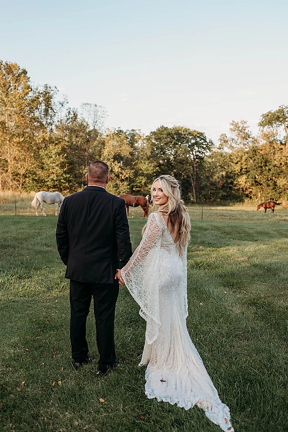 Couple portrait of newlyweds holding hands, bride looking back with a long train wedding dress in a grassy field near horses and fence