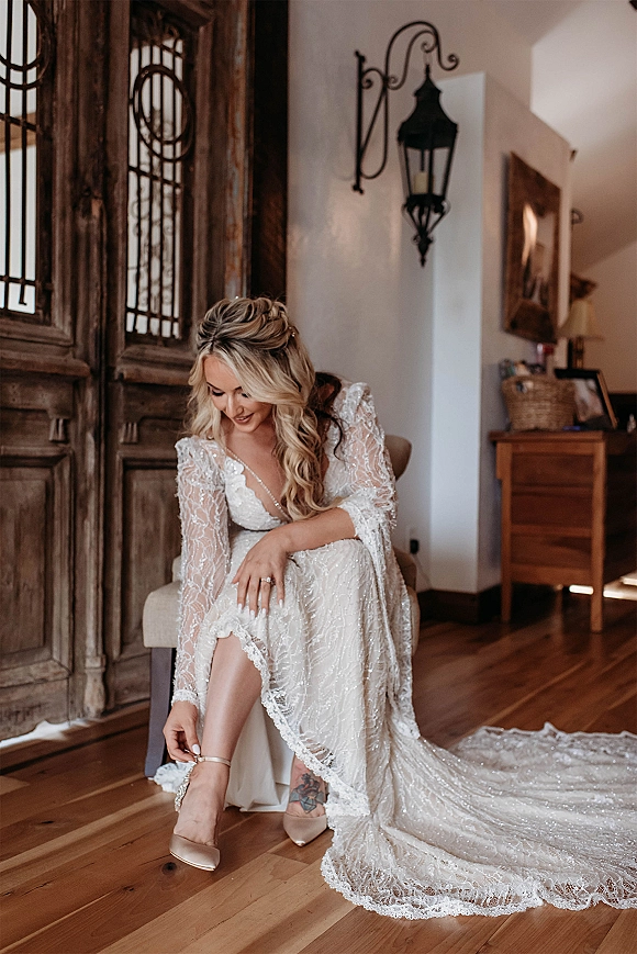 Bridal getting ready bride adjusts nude ankle-strap heel as lace long-sleeve gown train falls near wooden double doors indoors