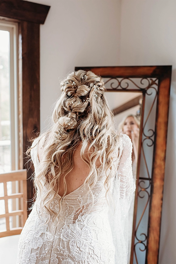 Bridal hair styled half up with loose curls, secured with pearl pins, seen from behind in mirror by soft window light