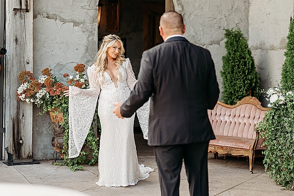 First look moment as bride in a lace wedding dress with arms open faces groom in black suit by a plaster wall doorway and greenery
