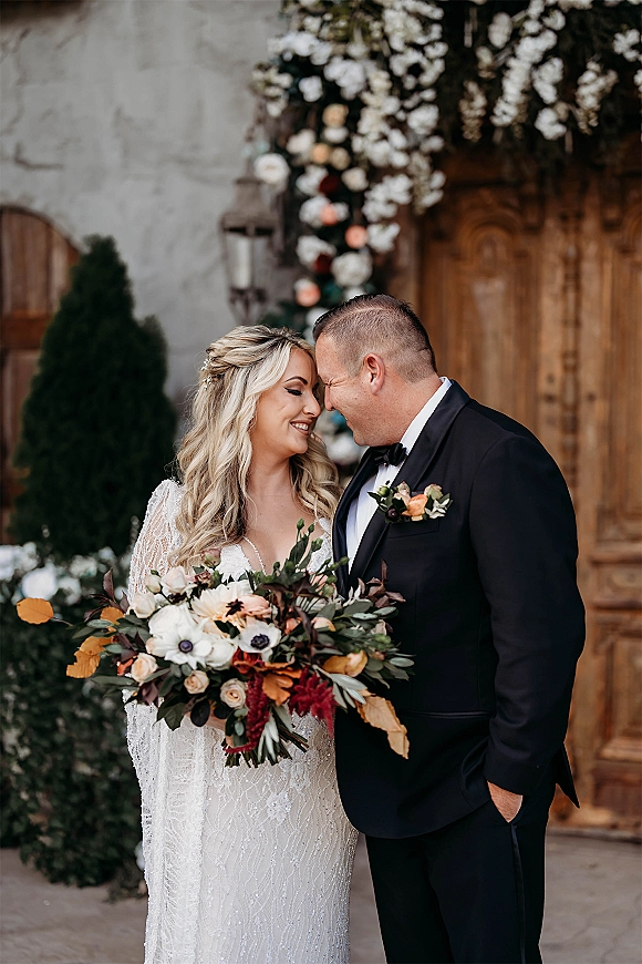 Couple portrait of bride and groom touching foreheads, bride holding a bouquet, posed by a rustic wooden door and stone wall greenery