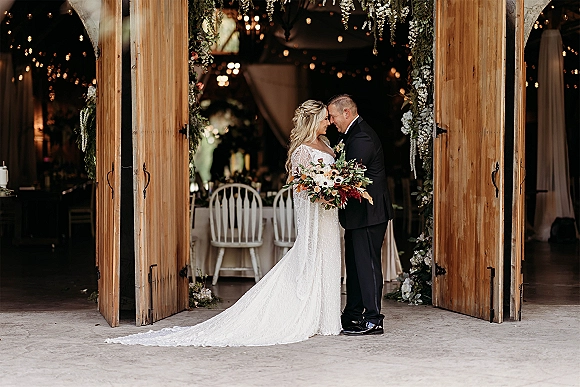 Couple portrait of bride and groom embrace with bouquet, lace dress train and tuxedo under floral arch by barn doors and string lights