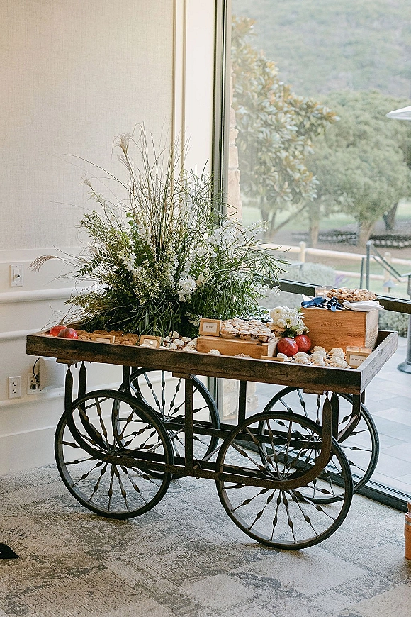 Dessert table on a wedding dessert cart with cookies and assorted pastries, wooden crates, candles, and florals by a large window