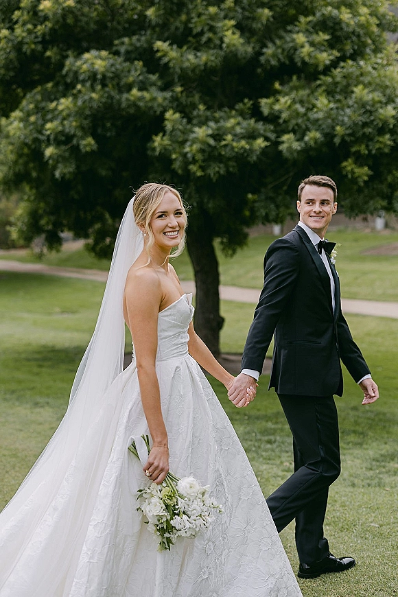 Couple portrait of bride and groom holding hands, her long veil and white bouquet flowing as they walk on a green lawn path