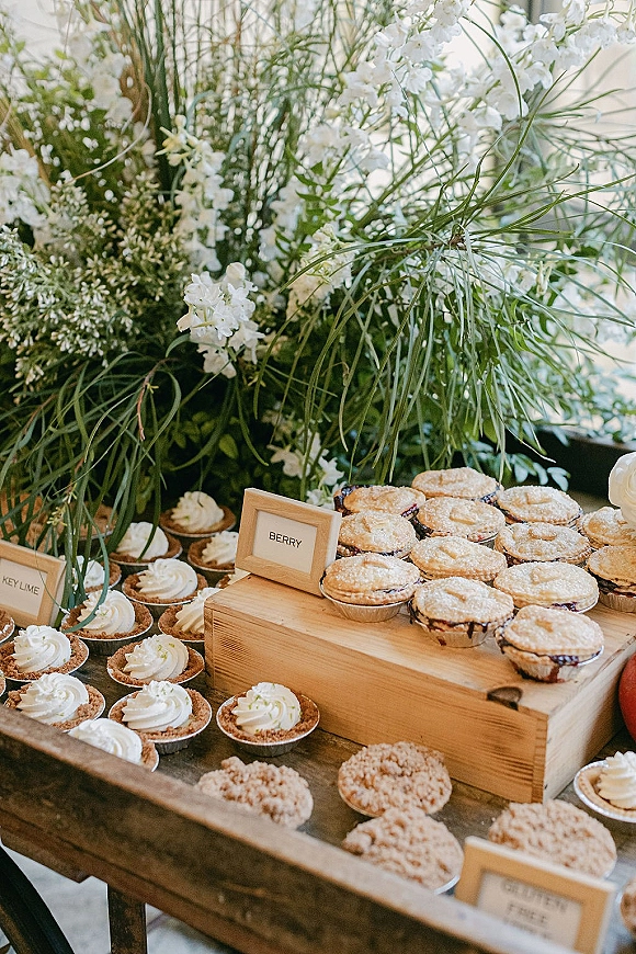 Wedding dessert table with mini pie dessert bar featuring tarts and cupcakes on wooden risers, labeled trays, and greenery by window light