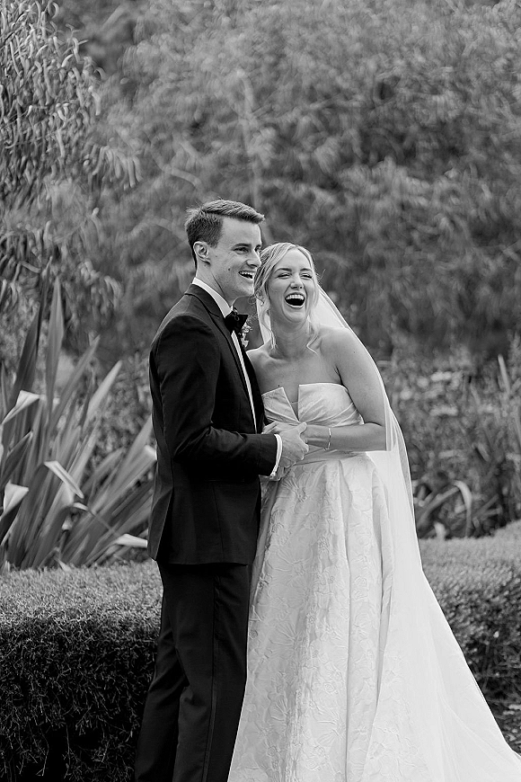 Wedding couple portrait of bride and groom laughing, holding hands in a black and white garden scene with veil, tuxedo, and greenery backdrop