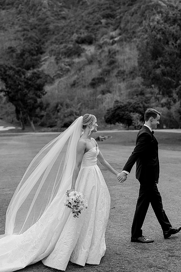 Couple portrait in a black and white wedding portrait style, bride and groom holding hands on a lawn with trees and hillside behind