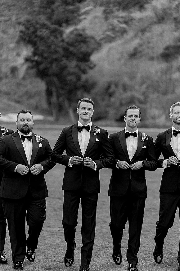 Groomsmen portrait of men walking in black tuxedos with bow ties and boutonnieres across an outdoor lawn with trees behind