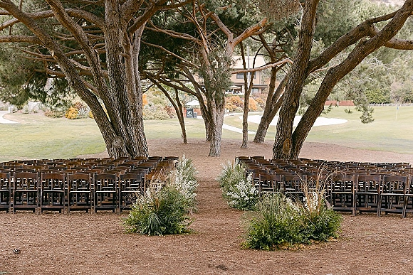 Ceremony setup with wood folding chairs and grounded aisle florals of white flowers and greenery on a dirt path beneath large trees