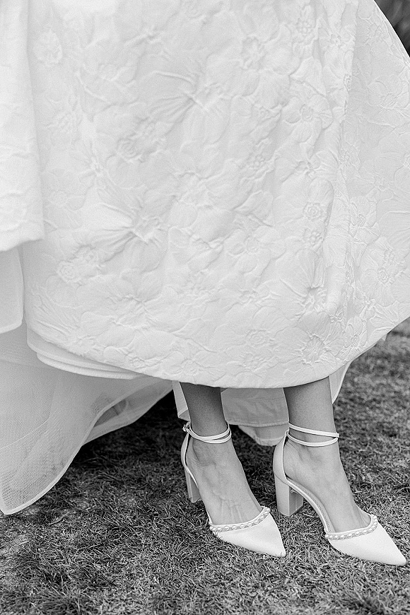 Wedding shoes with pearl wedding shoes trim, pointed-toe ankle-strap heels beside a bridal gown hem on a grass lawn