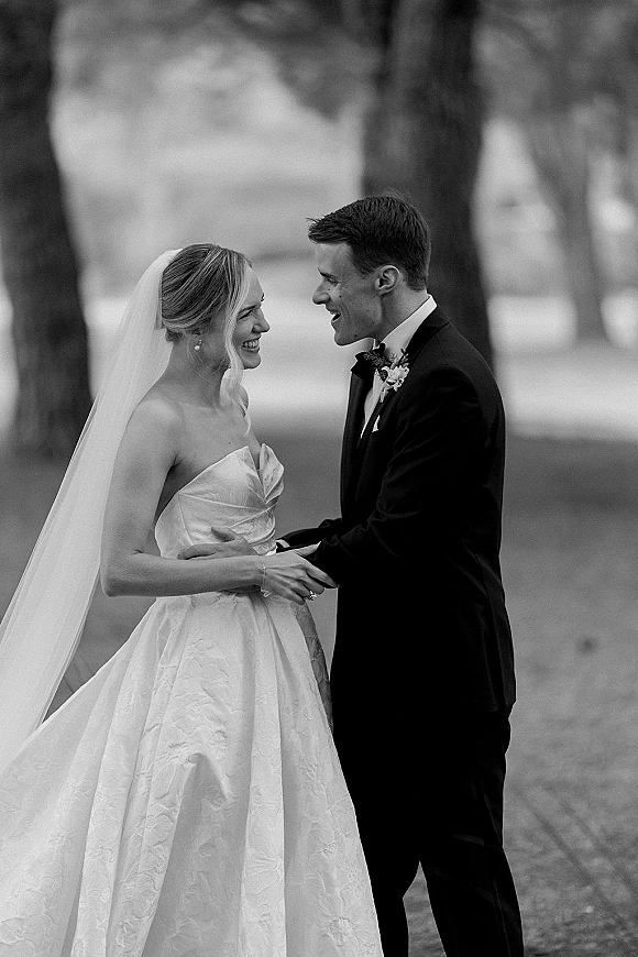 Couple portrait in a black and white wedding portrait, bride and groom holding hands and laughing on a tree-lined outdoor path with veil