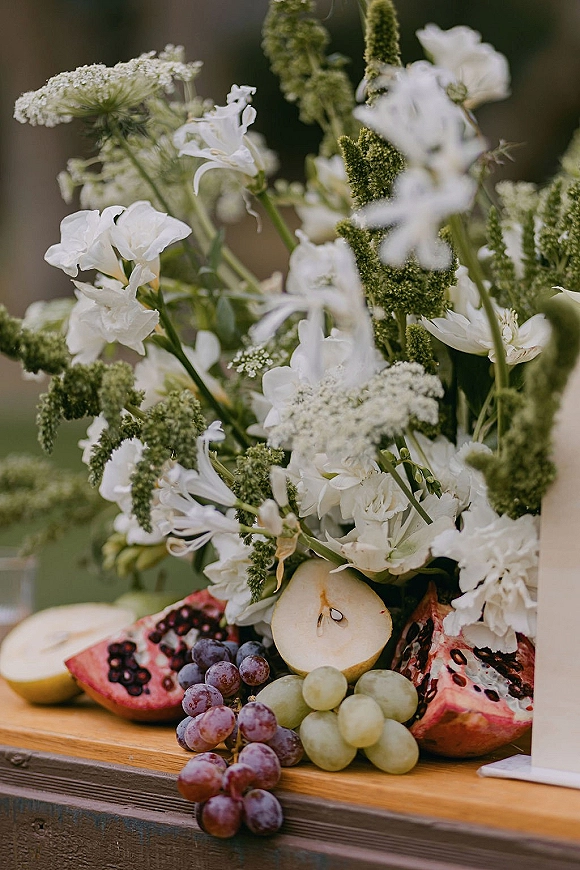 Wedding grazing table with a wedding fruit display of grapes, pear, and pomegranate beside white florals and greenery on a rustic wooden board outdoors