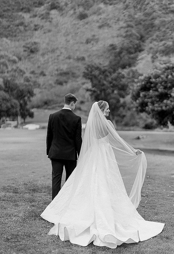 Couple portrait in a black and white wedding portrait, bride and groom walking away across a grassy field with hills and trees behind
