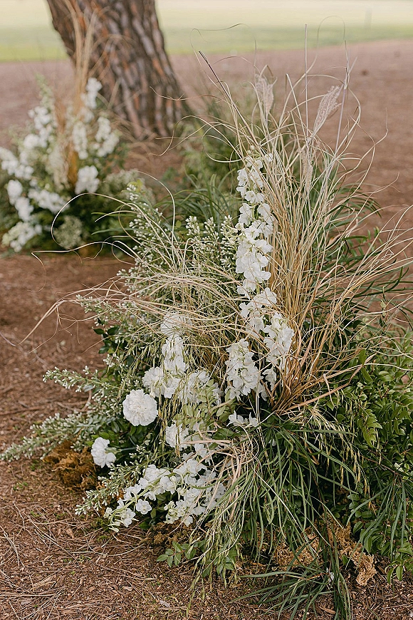Ceremony aisle florals with white flowers and greenery nestled at the base of a tree along a dirt path, accented with ornamental grasses