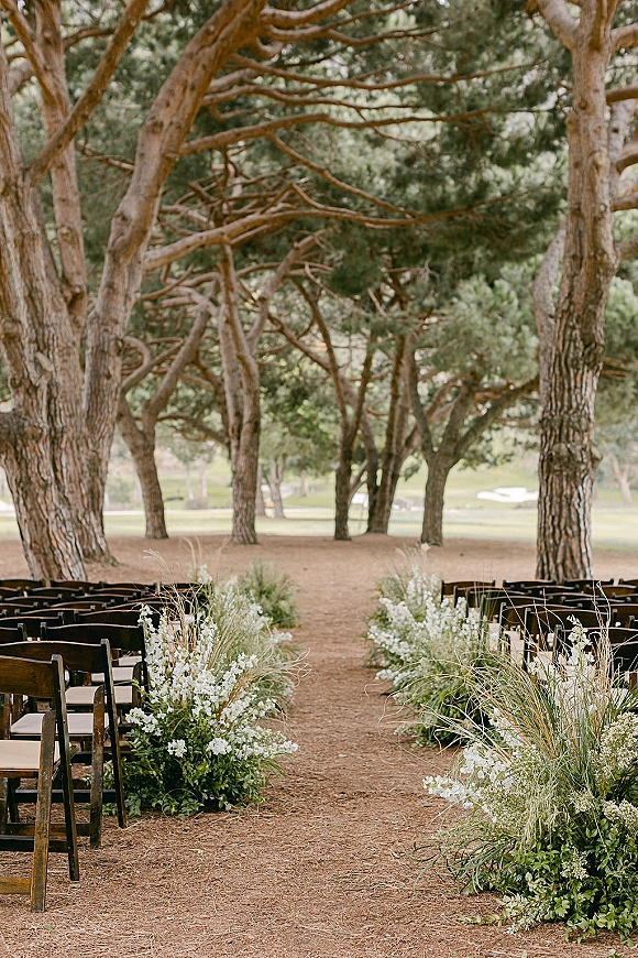 Ceremony aisle decor with outdoor wedding ceremony setup, wooden chairs and low white flower greenery clusters along a dirt aisle in pine forest