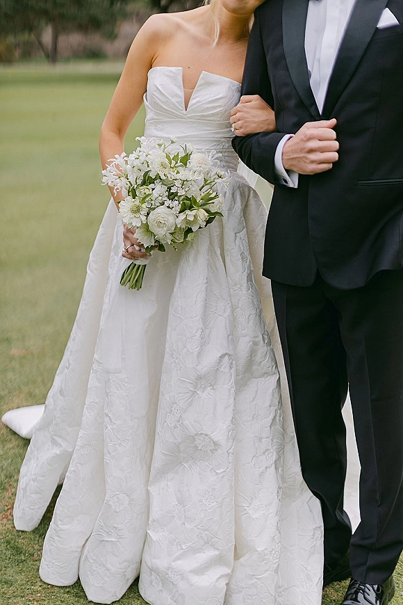 Couple portrait of bride and groom portrait, her strapless satin gown and white bouquet beside his tuxedo on a lawn by a stone wall