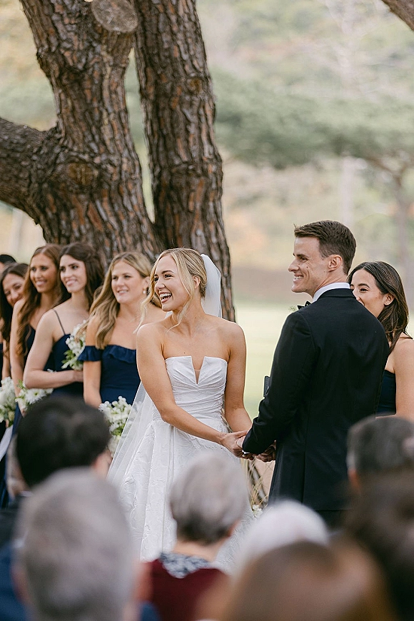 Ceremony moment as bride and groom holding hands, bride in strapless dress and veil with bridesmaids holding bouquets on a lawn with guests seated