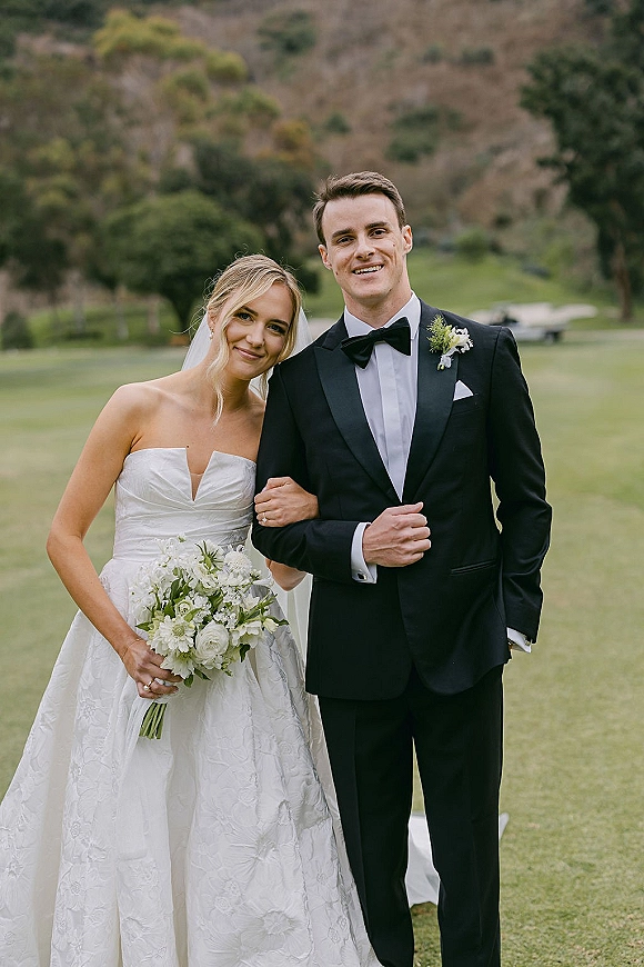 Couple portrait of bride in veil holding a white bouquet beside groom in black tuxedo on a green lawn with trees and hills backdrop